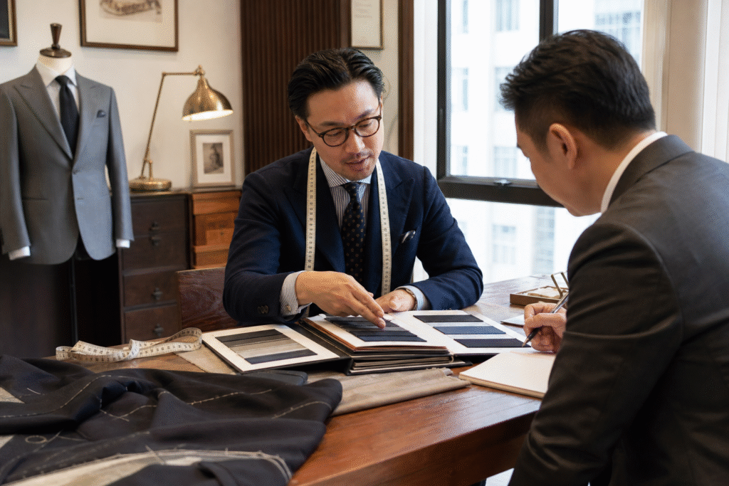 香港裁縫店內師傅向客人講解訂造西裝報價，桌上有布辦、量尺和西裝樣本，示範 bespoke 與 MTM 價錢差異